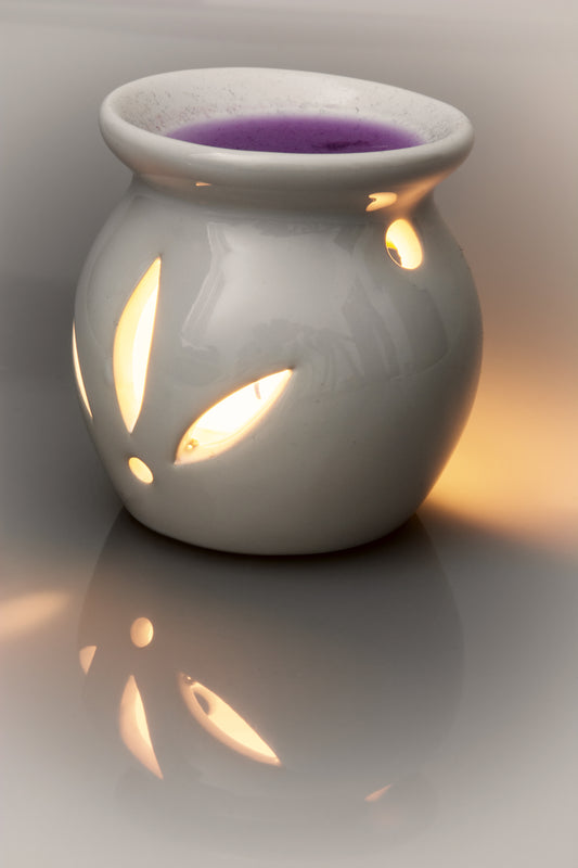 A white ceramic wax warmer with leaf-shaped cutouts, glowing warm from within, holding a purple wax melt, set against a dark gray gradient background with a soft reflection below.
