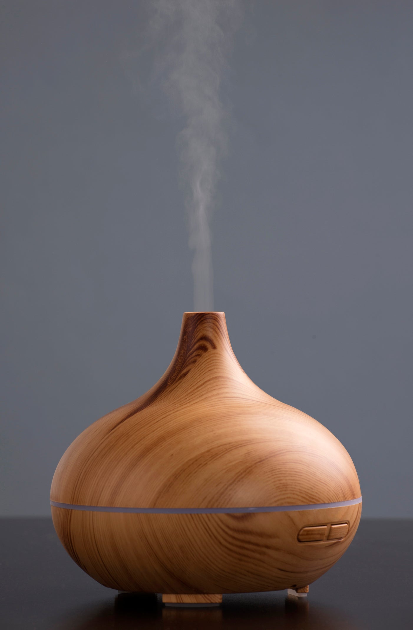A wooden essential oil diffuser with a rounded bulbous body and a small spout from which white mist is emitting, set against a dark gray background.