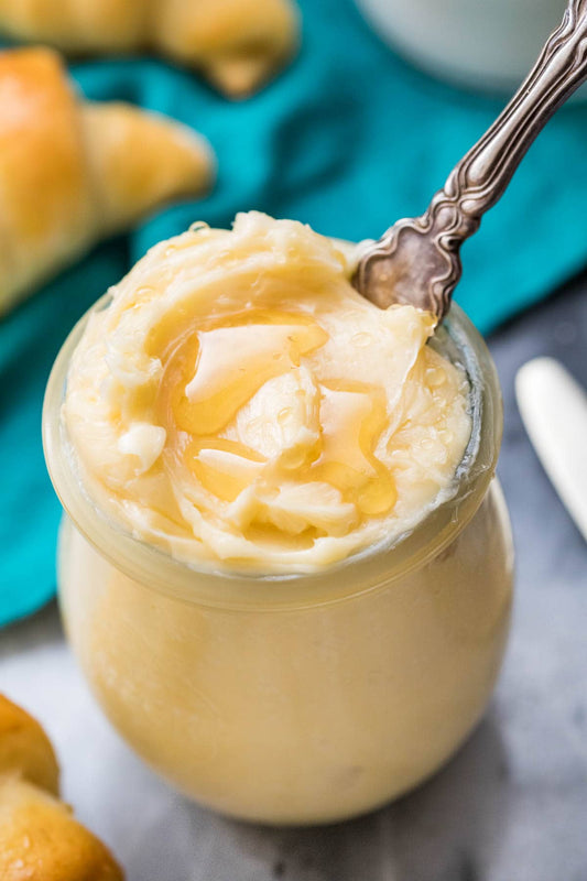 A close-up of a creamy pale yellow dessert or spread in a small glass jar with a spoon, topped with a drizzle of honey, with bread rolls blurred in the background on a teal cloth.
