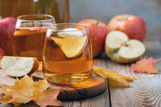 A glass of maple bourbon cocktail with an apple slice, surrounded by apples and autumn leaves on a rustic wooden table.