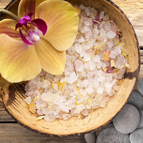 A bamboo bowl filled with white crystals, resembling sea salt, and adorned with the petals of an orchid, placed on a wooden surface with smooth rocks beside it.