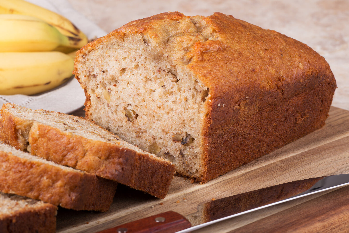A loaf of banana nut bread on a wooden cutting board with sliced bananas and a knife in the background.