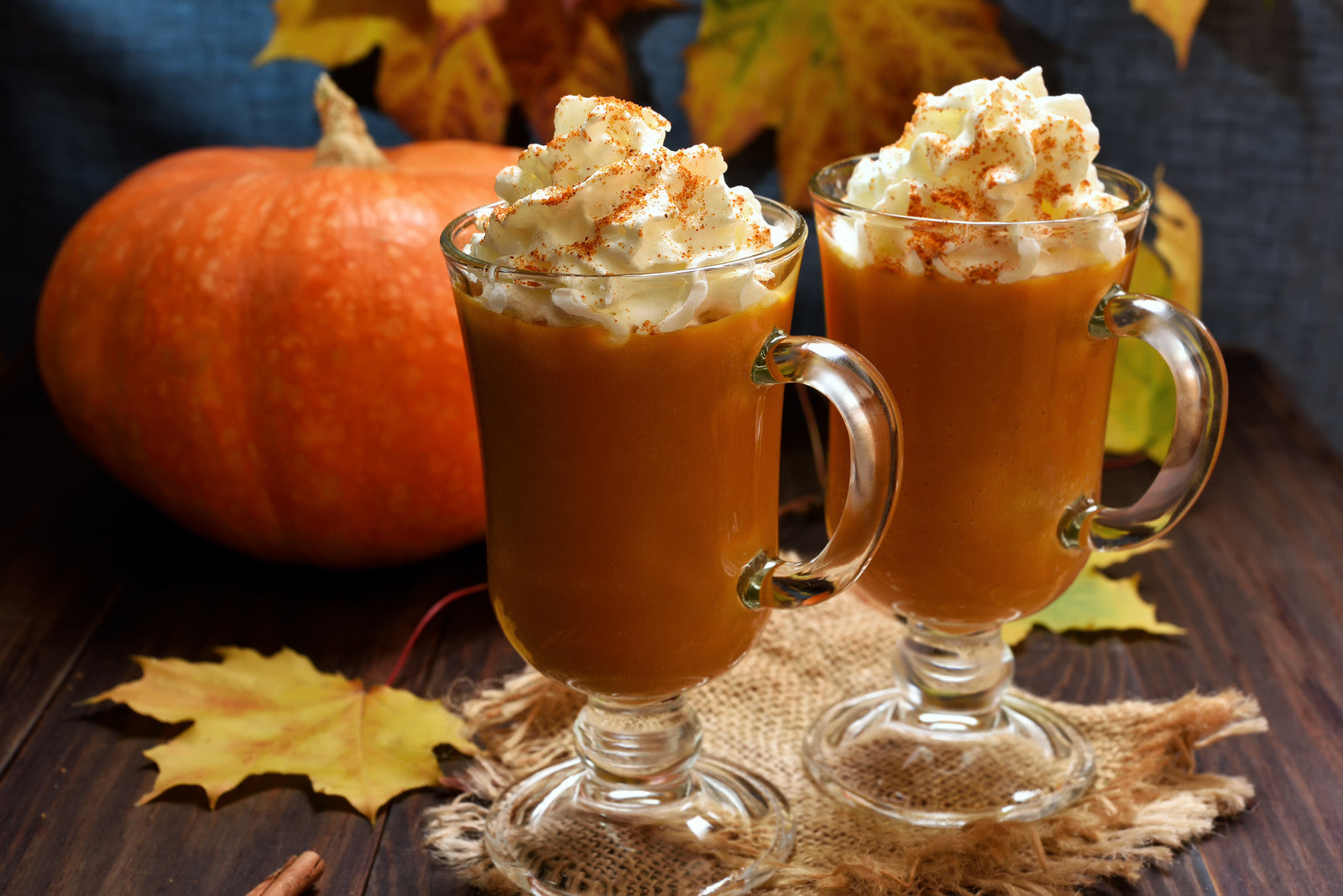 Two glasses of pumpkin spice simmering oil with whipped cream on top, garnished with nutmeg, on a burlap mat with a pumpkin and autumn leaves in the background.