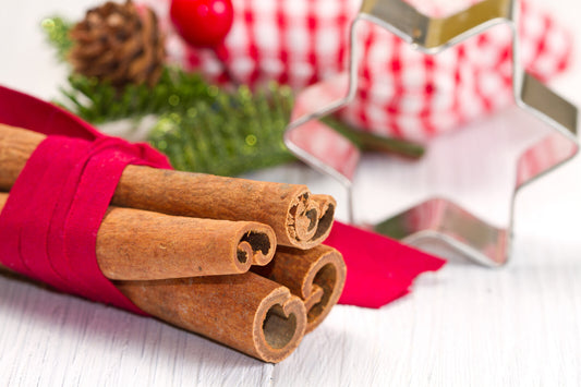 A bundle of cinnamon sticks tied with a red ribbon, with Christmas decorations in the background, including a pine cone and a star-shaped cutter.