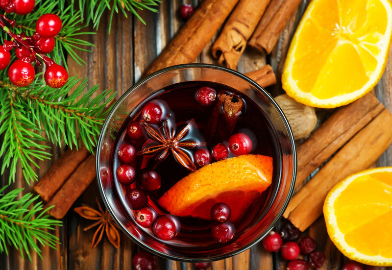 A glass bowl of cranberry simmering oil with orange slices and cinnamon sticks, surrounded by festive decorations like pine branches and more citrus fruit.