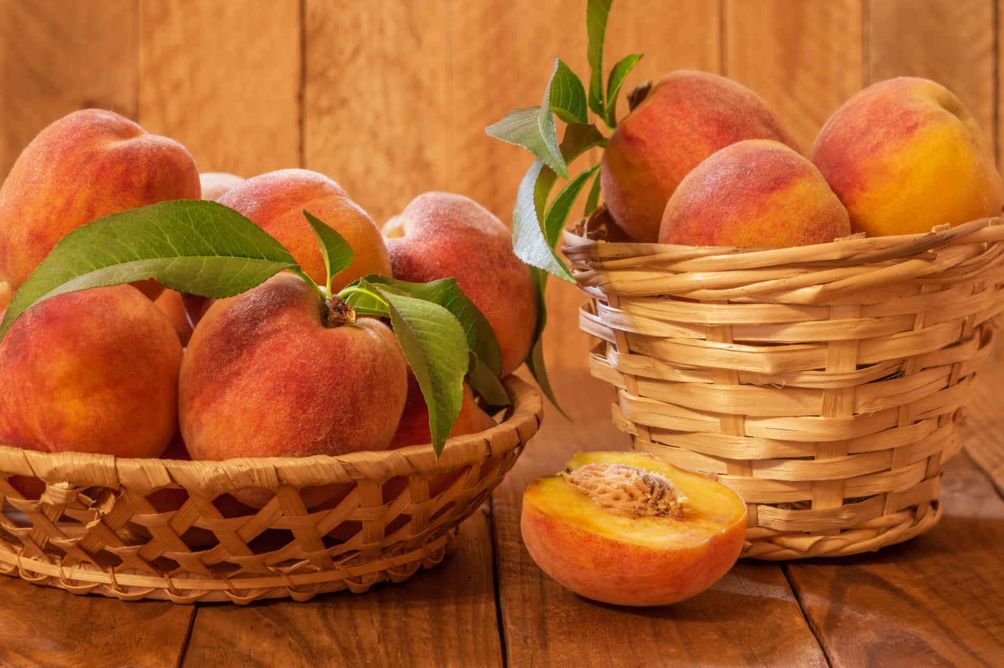 A wicker basket filled with fresh peaches on a wooden surface, with a few peaches placed outside the basket and a half-peach cut to show the inside.